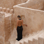 Model in a black halter maxi dress standing on a stone staircase in a sunlit courtyard.