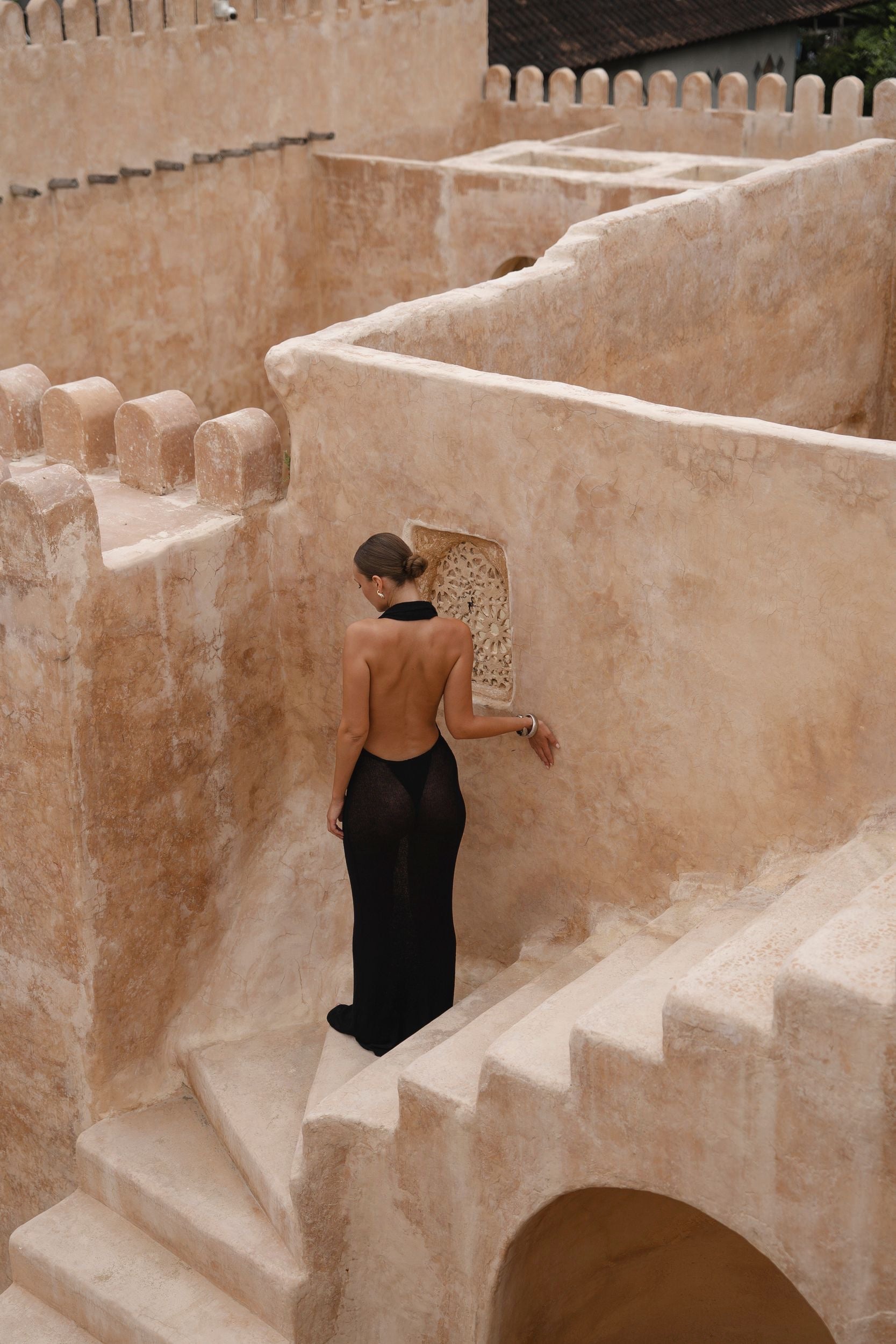 Model in a black halter maxi dress standing on a stone staircase in a sunlit courtyard.