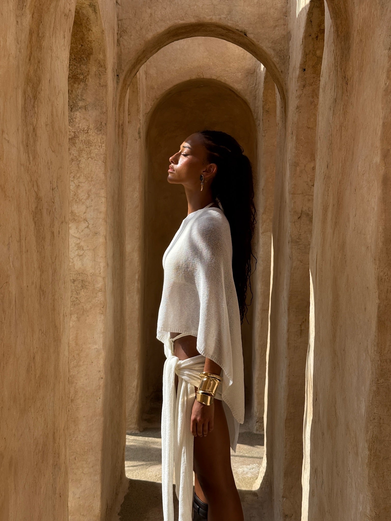 Model wearing an Ivory cape and matching sarong outfit standing in a sunlit archway of a beige building