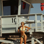 Model in a green pastel bikini standing next to a lifeguard tower on a beach.