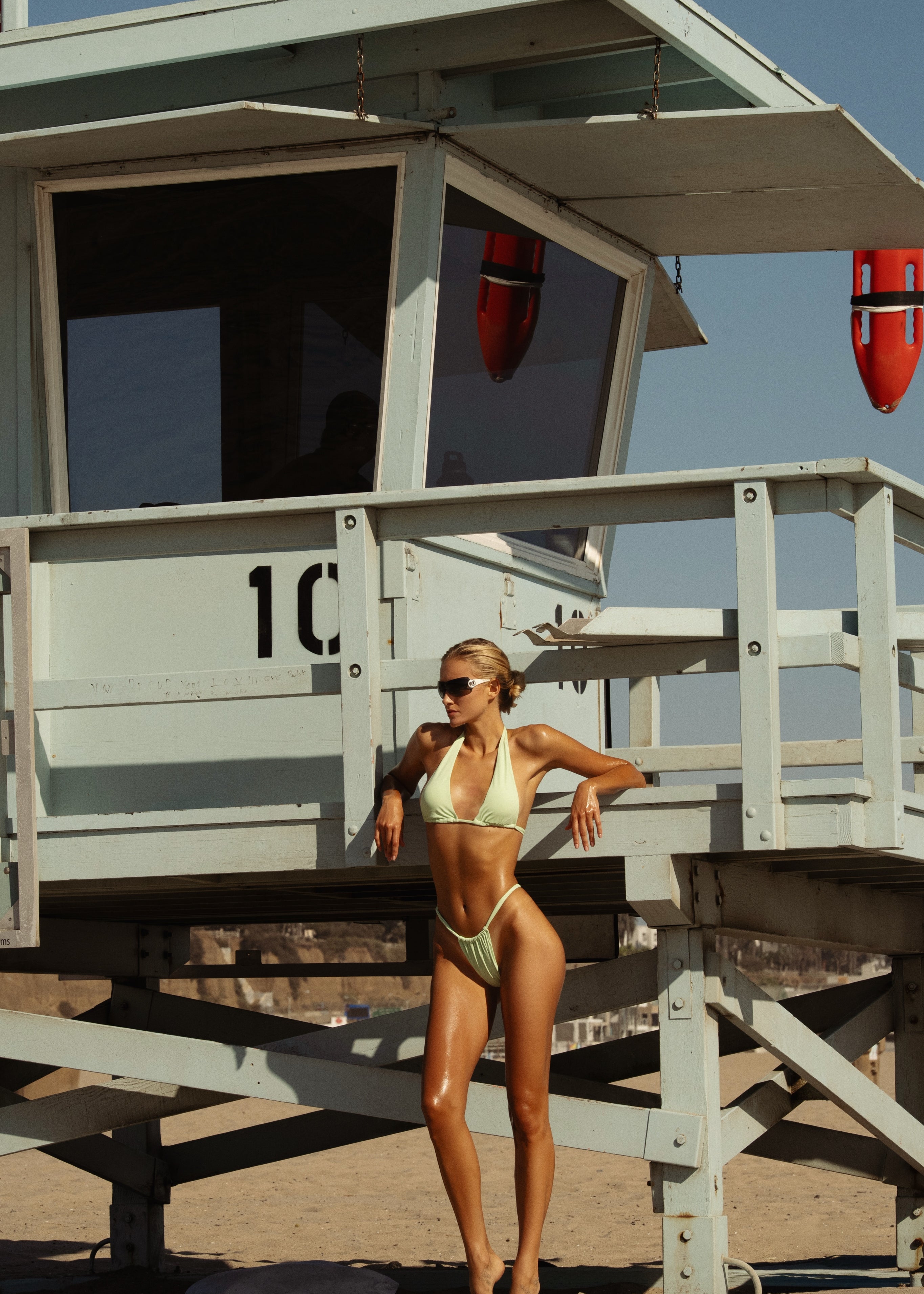 Model in a green pastel bikini standing next to a lifeguard tower on a beach.