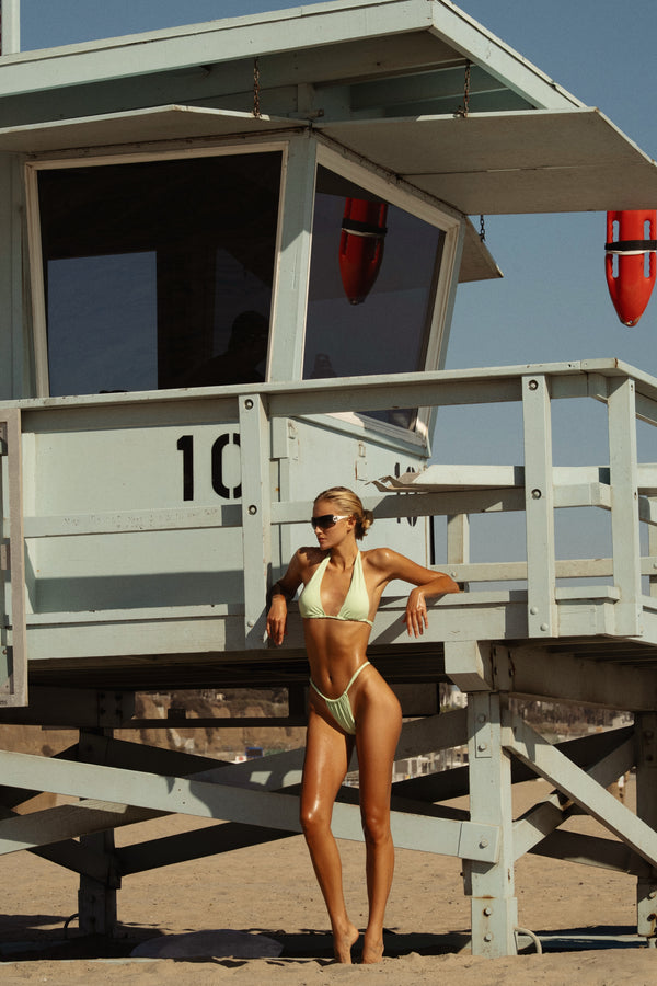 Model in a green pastel bikini standing next to a lifeguard tower on a beach.