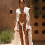 Model wearing an ivory sheer ruffled maxi cover-up with an ivory bikini, standing outdoors in sunlight against a textured wall with greenery.
