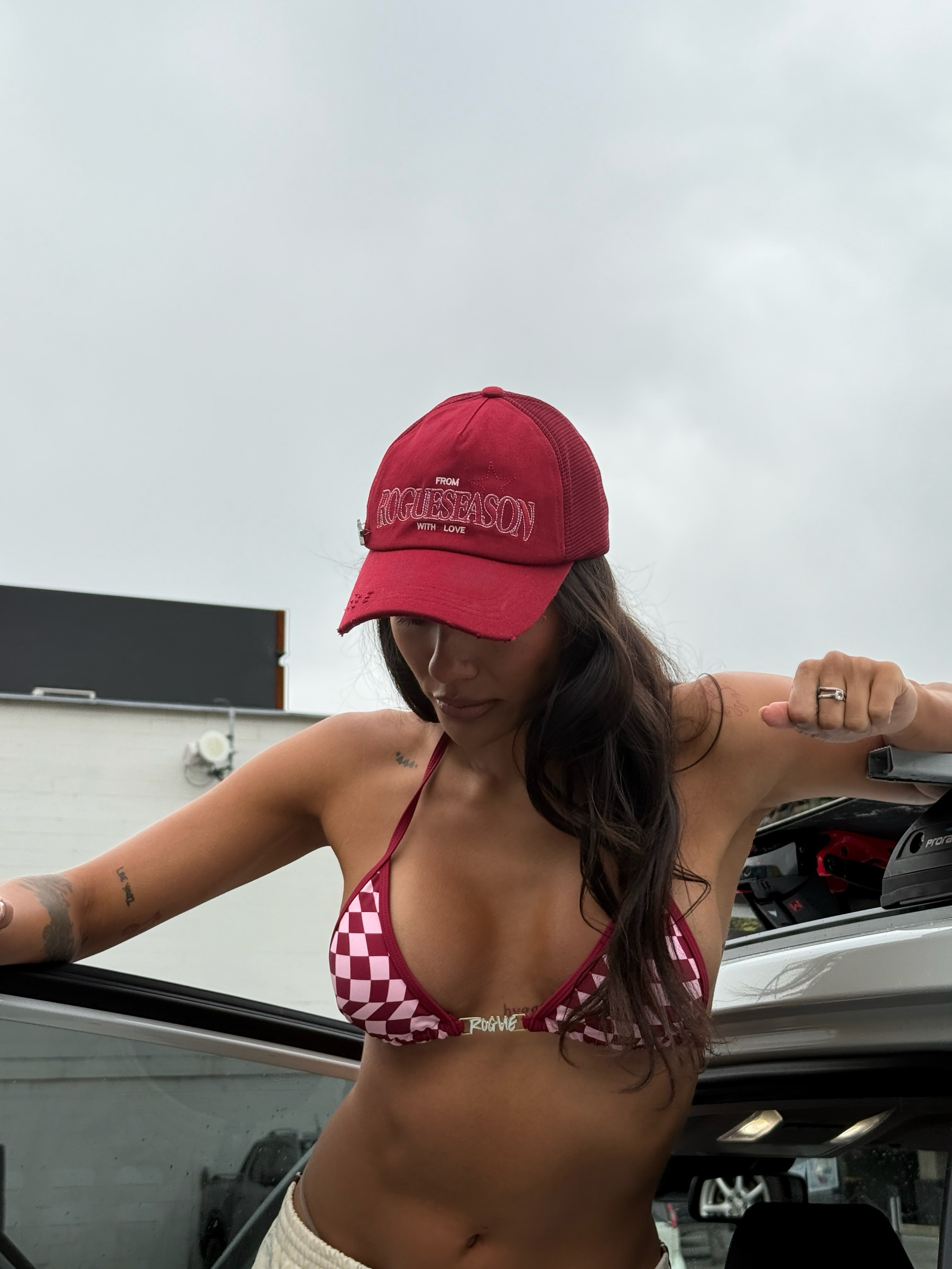 Model wearing a red cap and checkered bikini top standing next to a car.