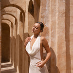 Cropped in shot of model wearing an Ivory halter neck maxi dress standing in a sunlit archway with sand-coloured walls.
