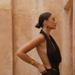 Side shot of model wearing a brown halter neck maxi dress with sand-coloured walls in the background.