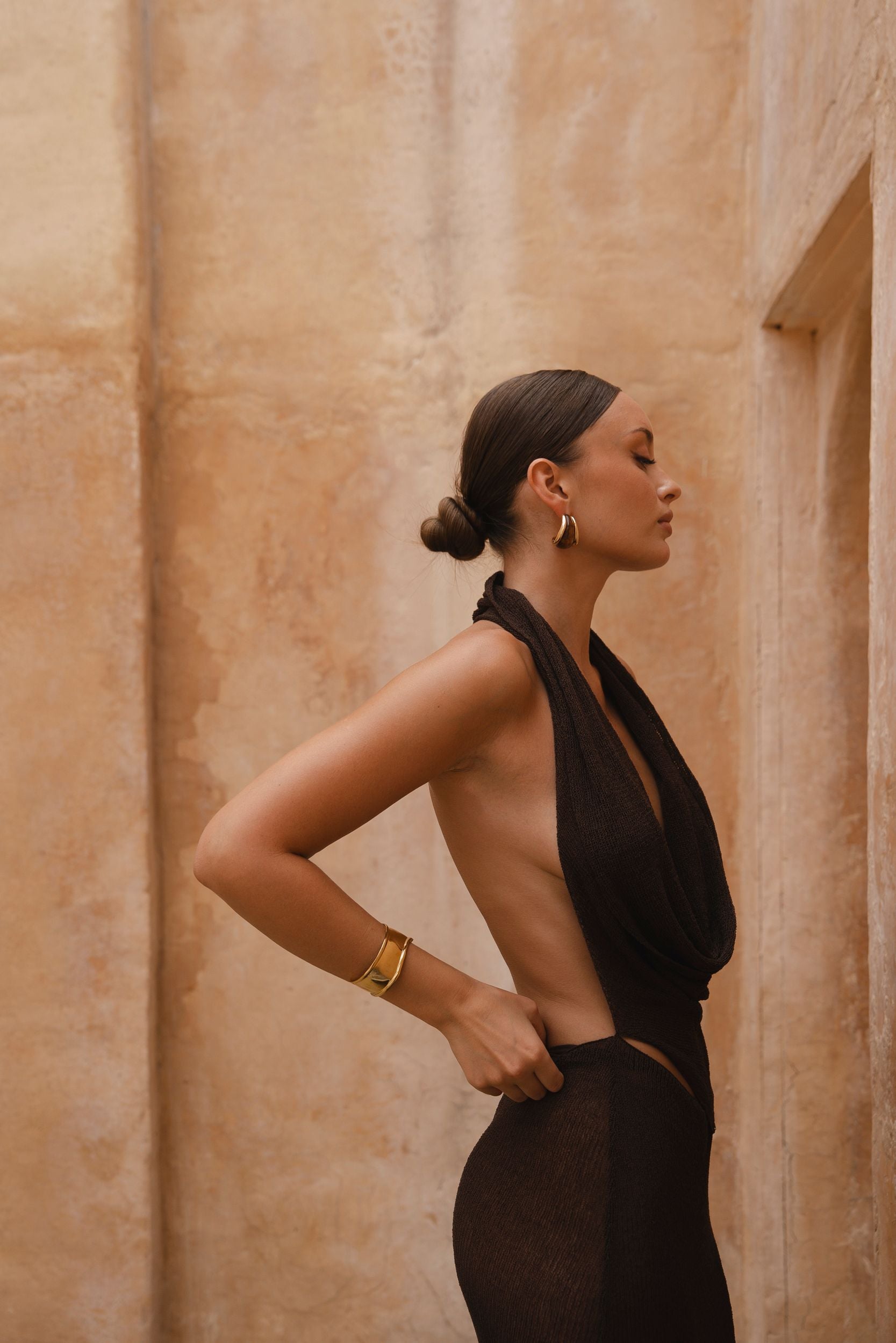 Side shot of model wearing a brown halter neck maxi dress with sand-coloured walls in the background.