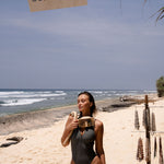 Model wearing a grey zip up one piece sleeveless swimsuit  with a cut-out back standing on a beach with ocean and sky in the background 