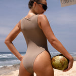 Back shot of model wearing a cinnamon zip up one piece swimsuit holding a coconut on a beach with a clear blue sky.