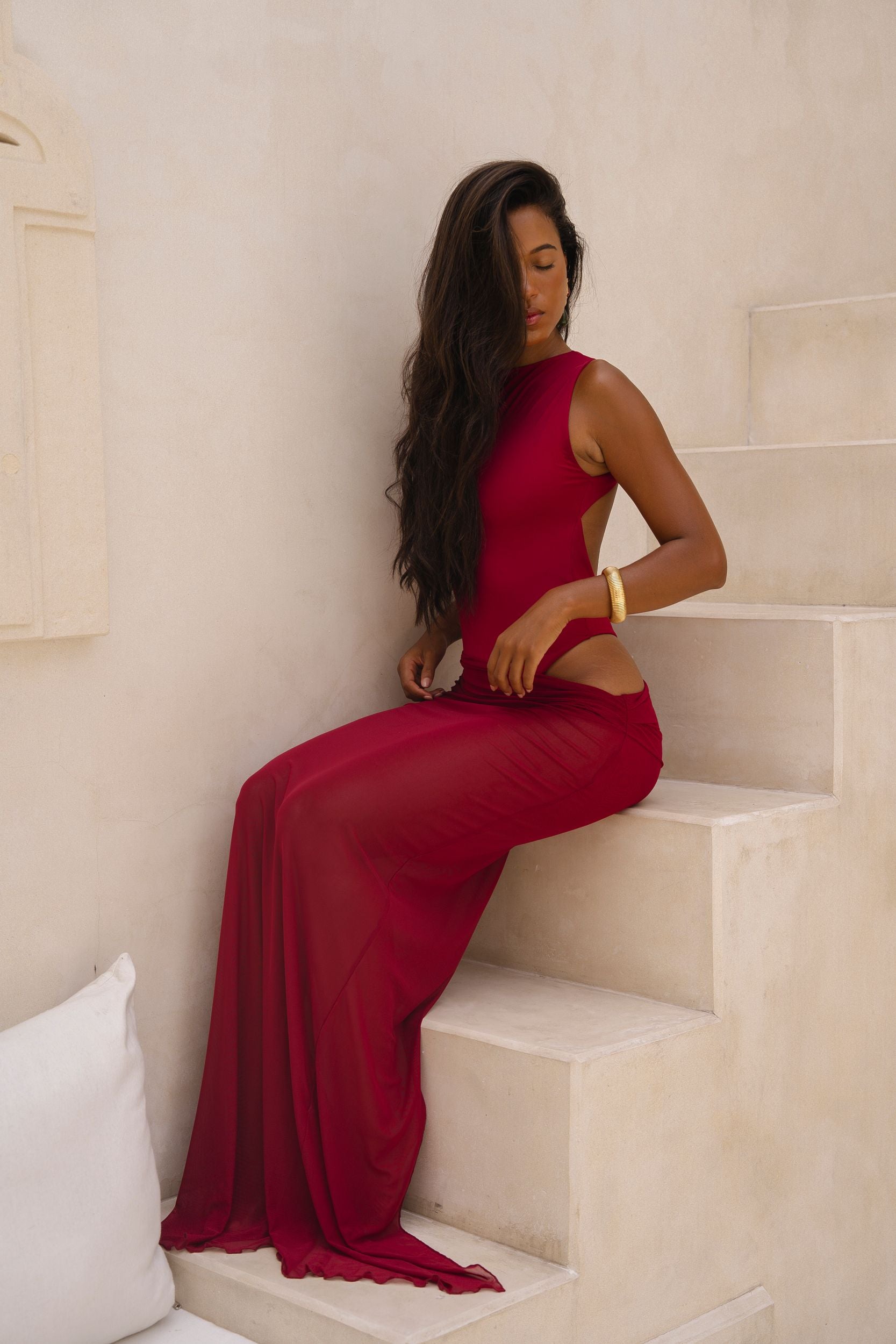 Model wearing a cherry Rogue Season one-piece swimsuit with side cut-out and matching sheer skirt, seated indoors in a neutral setting.