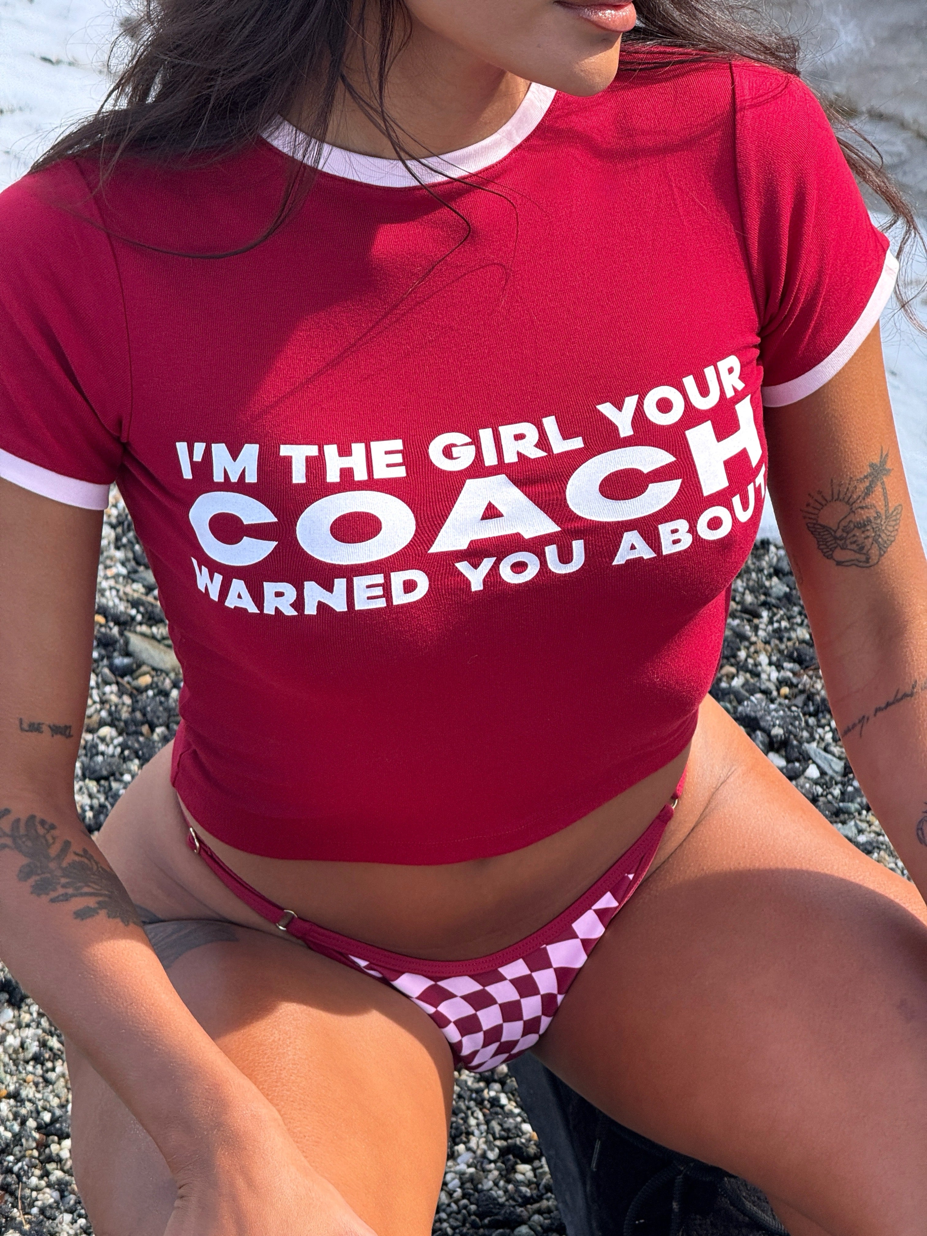 Close up shot of model wearing a cherry Rogue Season cropped t-shirt with printed text 'I'm the girl coach warned you about' and cherry checkered bikini bottoms.
