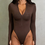 Close up of model wearing a brown long sleeve zip up one piece swimsuit next to a wall.