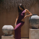Model in a purple outfit standing between two large stone vases against a thatched roof background.