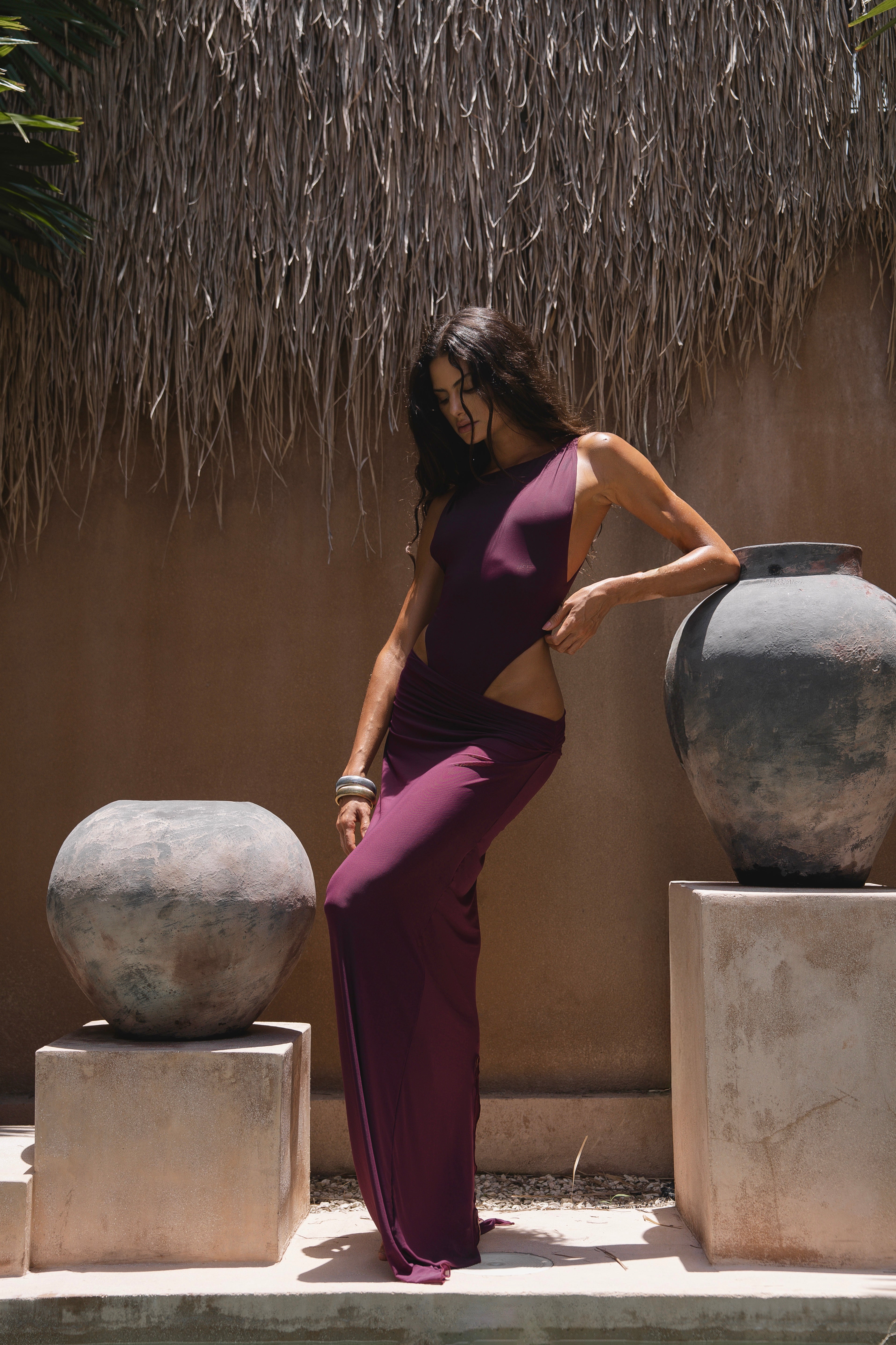 Model in a purple outfit standing between two large stone vases against a thatched roof background.