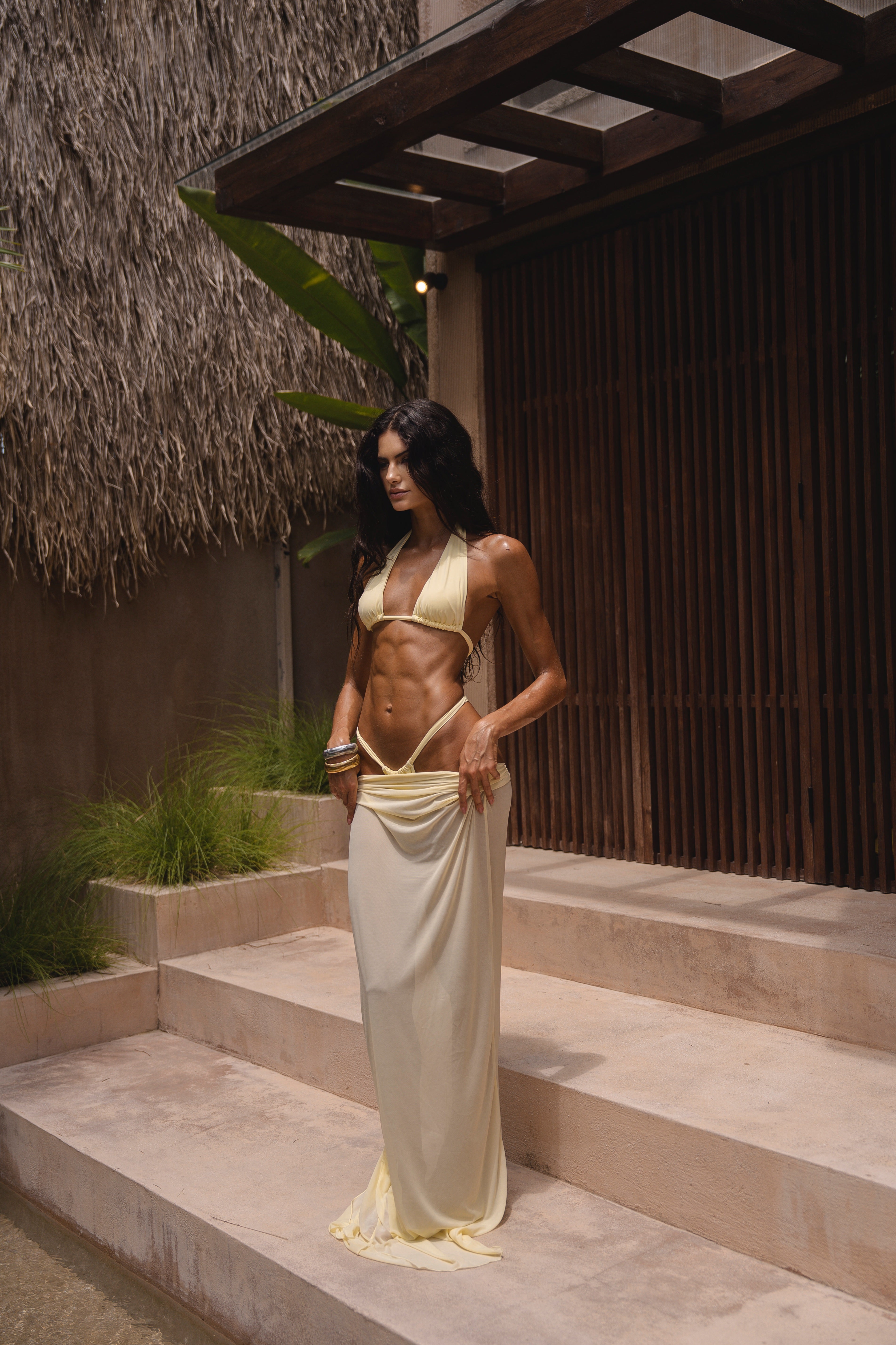 Model wearing a yellow swimsuit and matching sheer maxi skirt near a textured wall and standing on steps