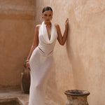 Model in an ivory maxi halter dress standing in a warm-toned stone setting with vases.