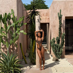 Model wearing a brown asymmetric cut-out backless swimsuit, standing in a tropical setting.
