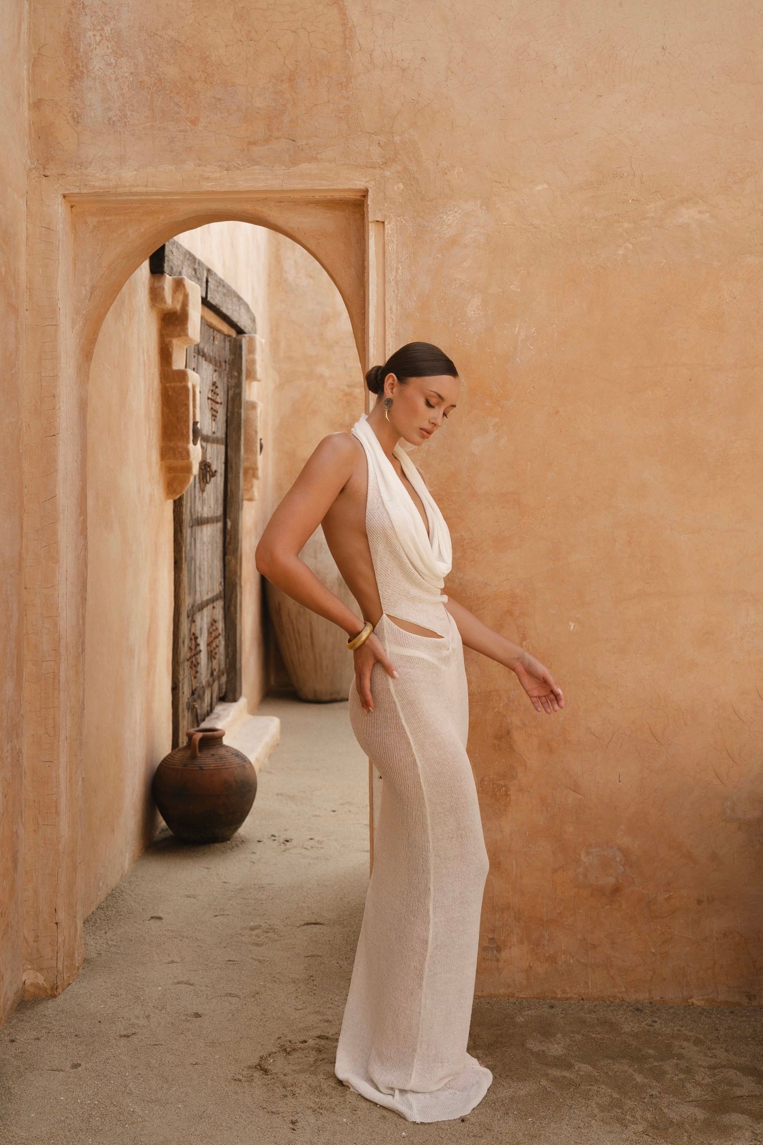 Side shot of model in a white dress standing in a sunlit archway with a warm-toned wall.