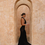 Model wearing a black halter neck maxi dress standing in a sunlit archway with sand-coloured walls.