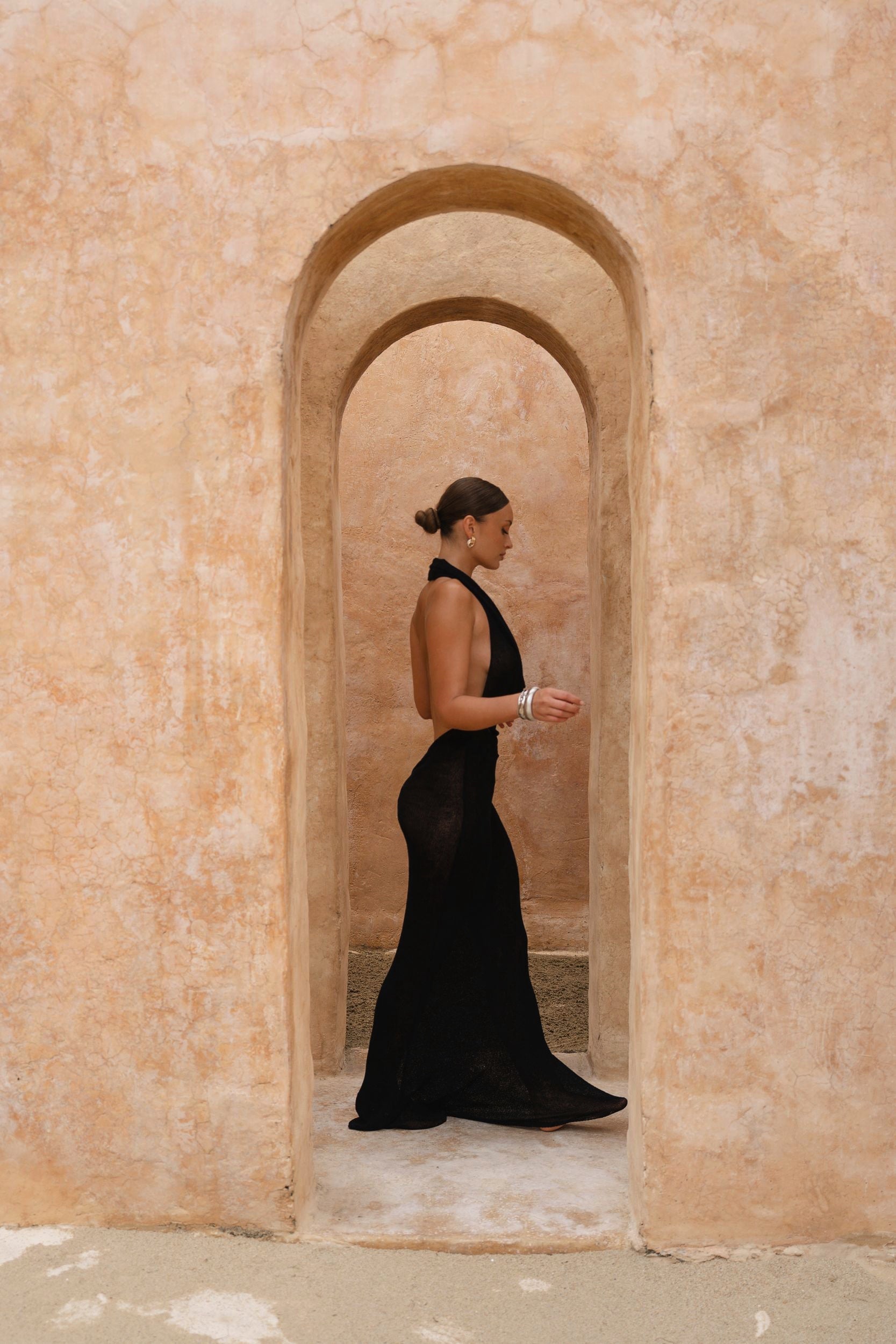 Model wearing a black halter neck maxi dress standing in a sunlit archway with sand-coloured walls.