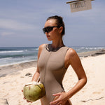 Model wearing a cinnamon coloured zip up one piece swimsuit holding a coconut on a beach with a clear blue sky.