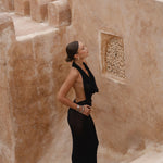 Model wearing a black halter dress standing on stone steps with a textured wall in the background