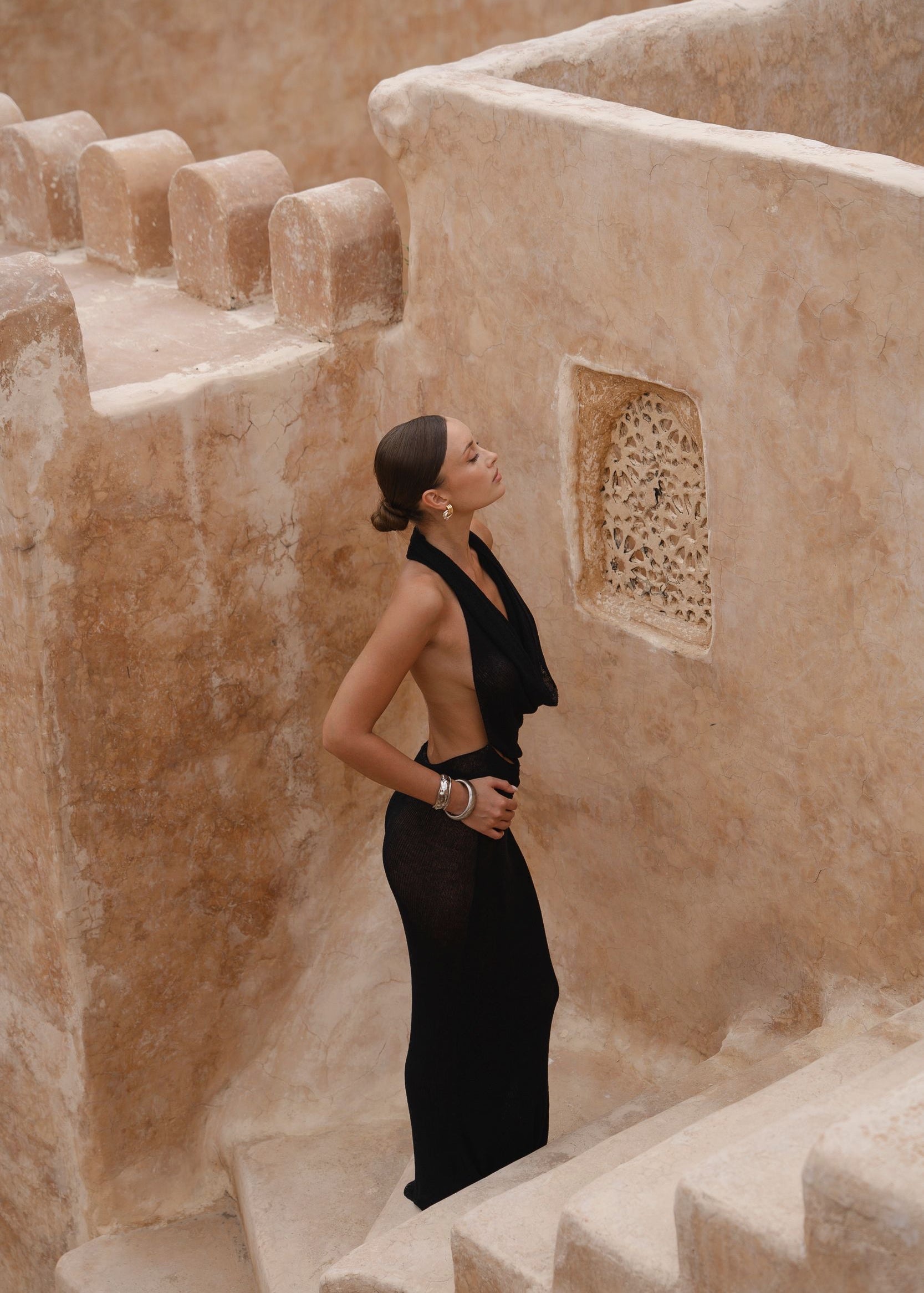 Model wearing a black halter dress standing on stone steps with a textured wall in the background
