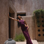 Model in a purple one piece swimsuit and sheer maxi skirt standing against a textured wall with architectural elements.