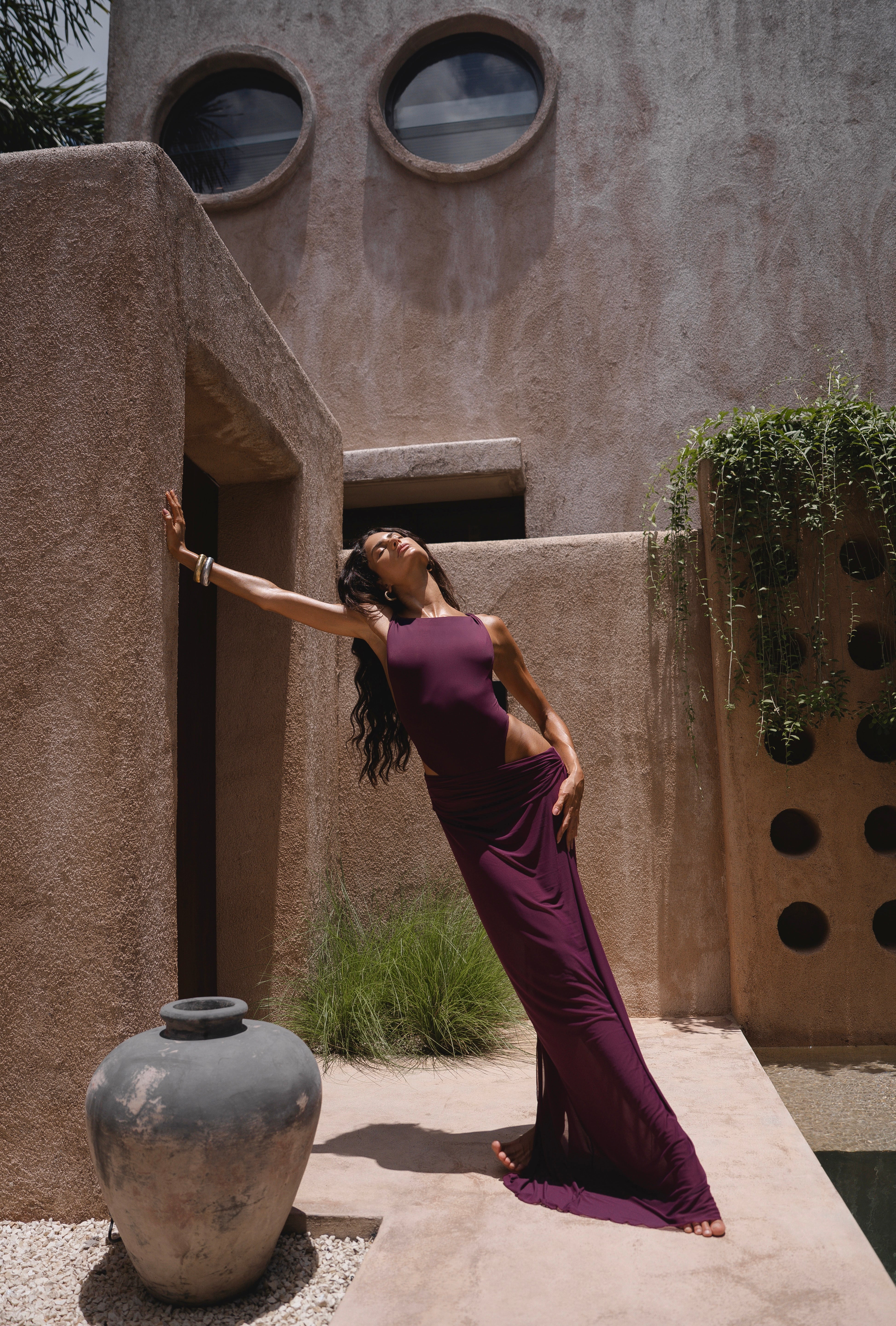 Model in a purple one piece swimsuit and sheer maxi skirt standing against a textured wall with architectural elements.