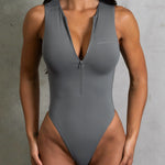 Very close up crop in detail of model wearing a charcoal coloured zip up one piece swimsuit in front of a grey wall.