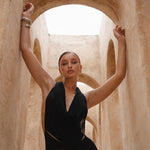 Model wearing a black halter neck dress standing in a sunlit archway with sand-coloured walls.