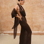 Side shot of model wearing a brown halter neck maxi dress standing near sand-coloured walls.