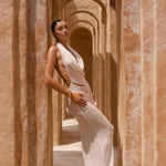 Model wearing an Ivory halter neck maxi dress standing in a sunlit archway with sand-coloured walls.