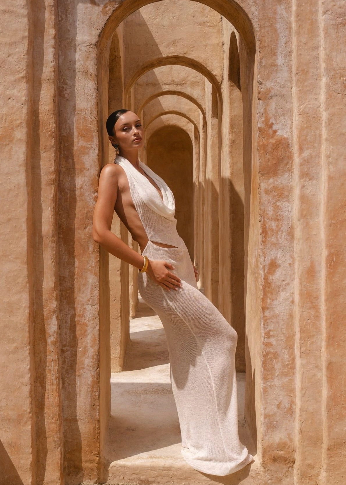 Model wearing an Ivory halter neck maxi dress standing in a sunlit archway with sand-coloured walls.