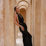 Model wearing a black halter neck maxi dress standing in a sunlit archway with sand-coloured walls.