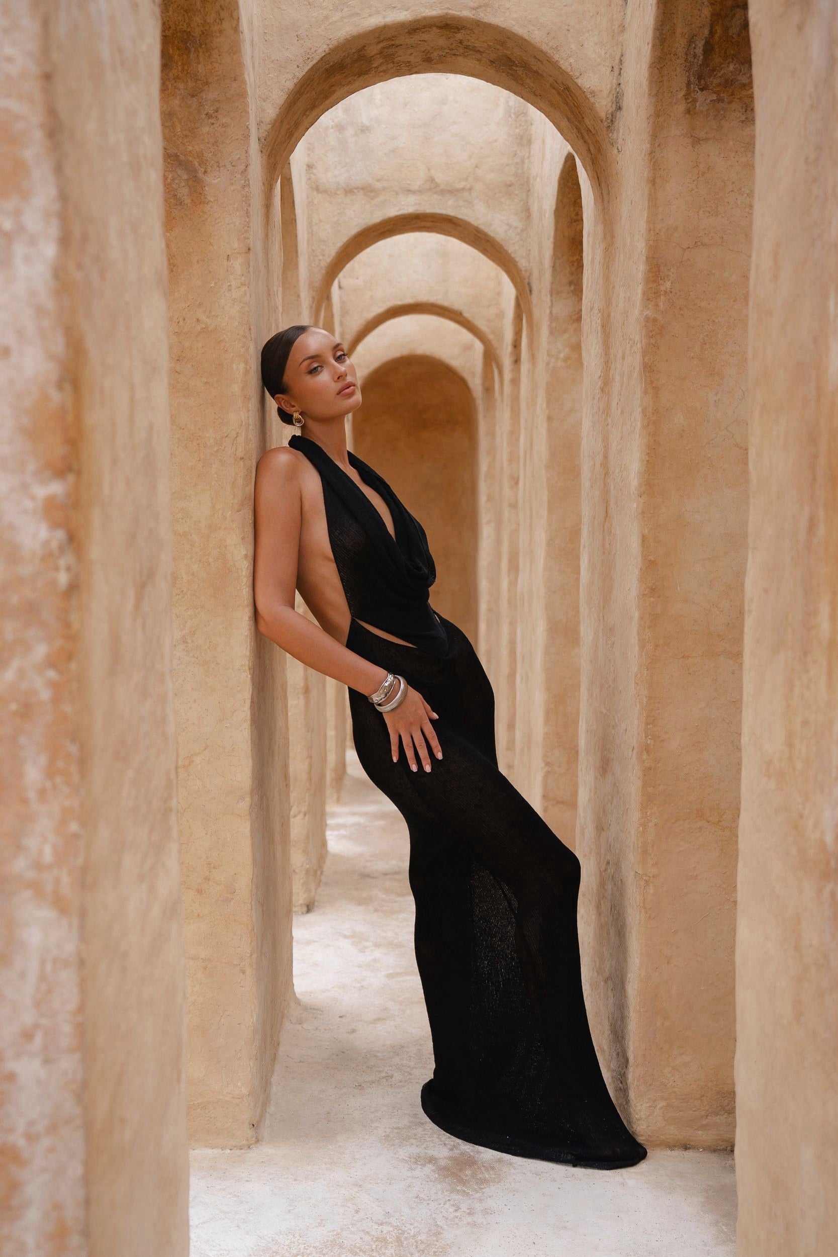 Model wearing a black halter neck maxi dress standing in a sunlit archway with sand-coloured walls.