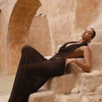 Model sitting and leaning back on stairs wearing a brown maxi halter dress.