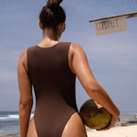 Back shot of model in a brown zip up swimsuit holding a coconut on a beach with ocean and sky in the background.