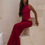 Model wearing a cherry Rogue Season one-piece swimsuit with side cut-out and matching sheer skirt, seated indoors in a neutral setting.