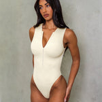 Model in a Ivory zip up swimsuit standing on in front of a grey wall.