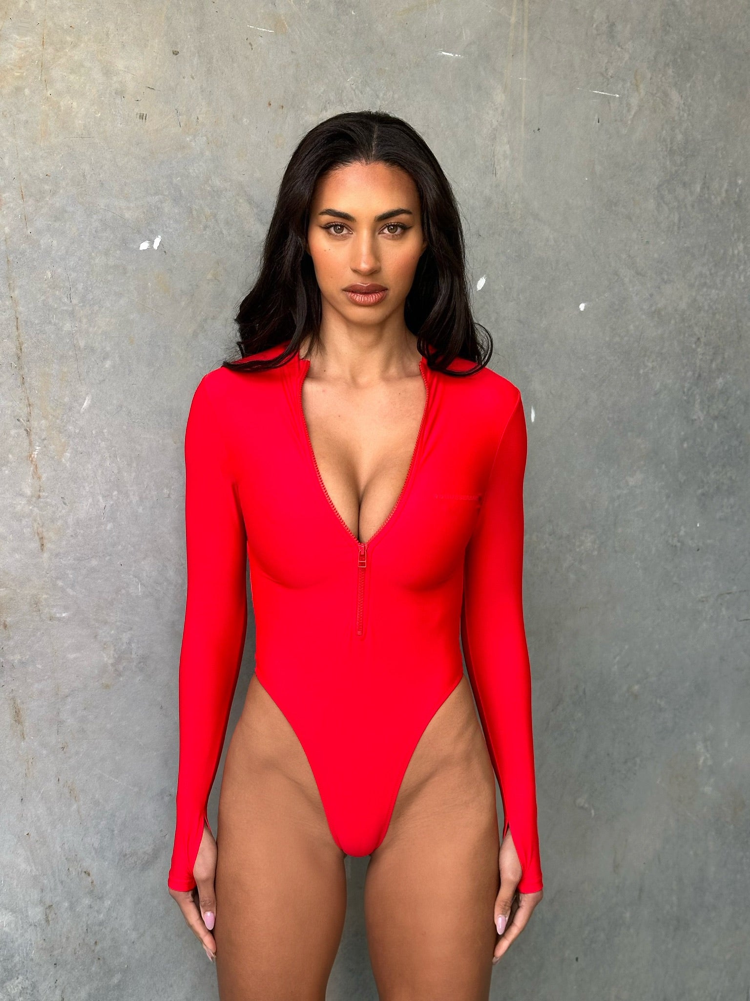 Model wearing a red long sleeve zip up one piece swimsuit in a studio with grey wall.