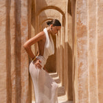 Model standing to the side wearing an Ivory halter neck maxi dress standing in a sunlit archway with sand-coloured walls.