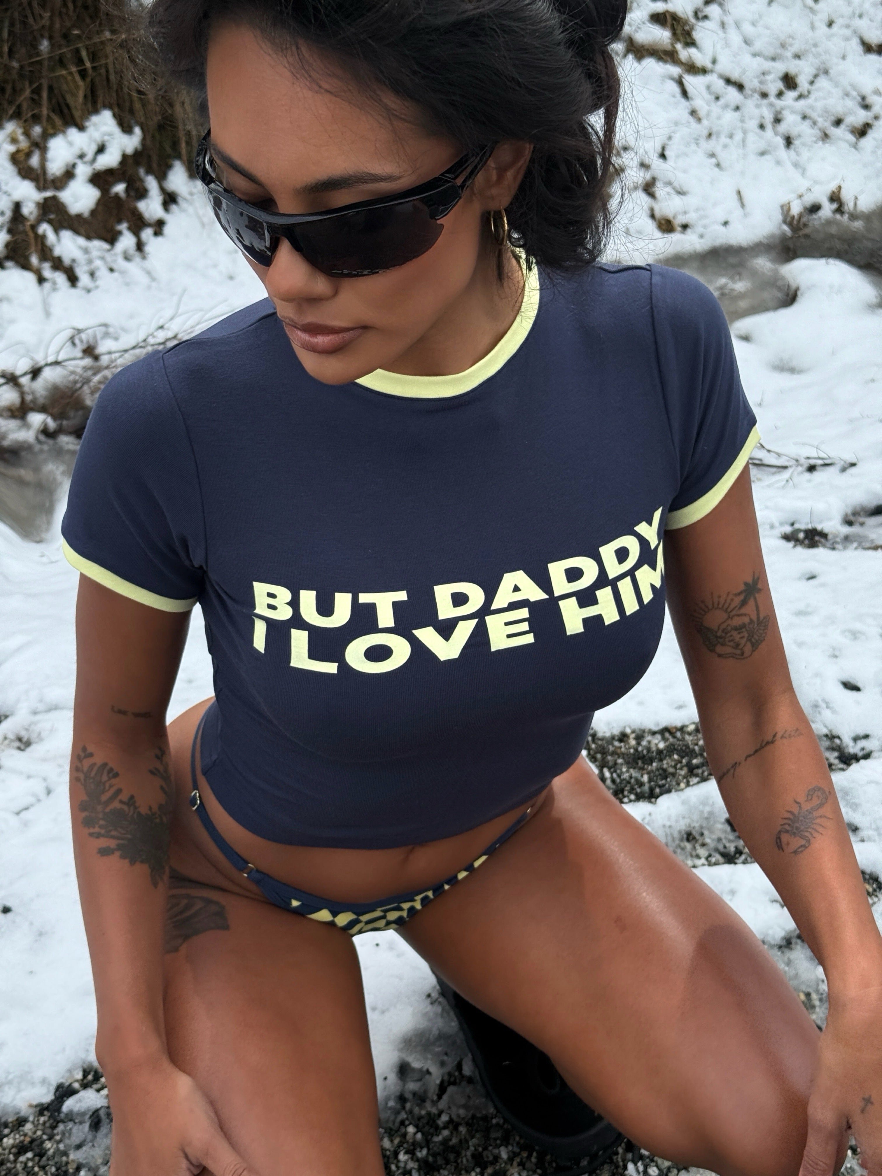 Model wearing a navy Rogue Season cropped t-shirt with printed text and navy and yellow checkered bikini bottoms, outdoors in a snowy environment.