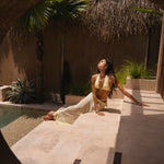 Woman in a yellow bikini and matching sheer maxi skirt sitting by a pool with palm trees and a thatched roof in the background