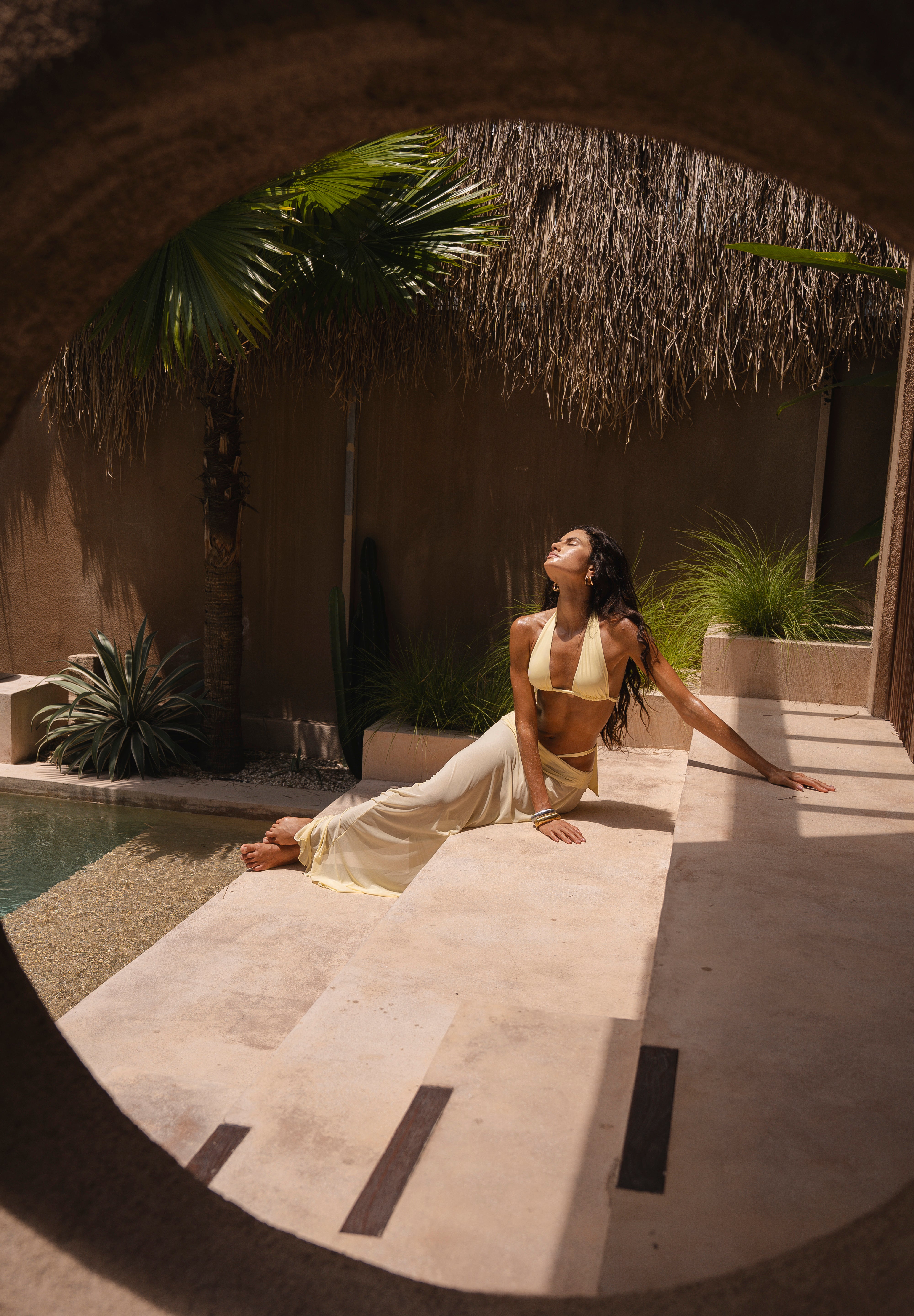 Woman in a yellow bikini and matching sheer maxi skirt sitting by a pool with palm trees and a thatched roof in the background