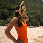 Model wearing a orange zip up one piece sleeveless swimsuit with a cut-out back standing on a beach with snorkelling goggles.