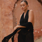 Model wearing a black halter neck maxi dress standing in a sunlit archway with sand-coloured walls.