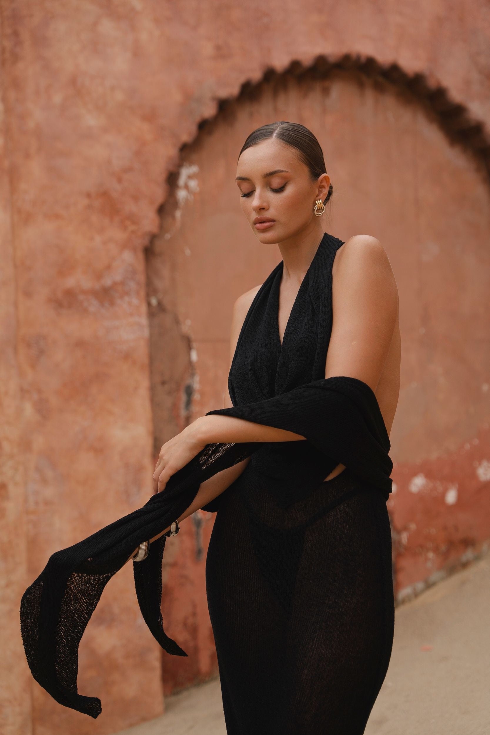 Model wearing a black halter neck maxi dress standing in a sunlit archway with sand-coloured walls.