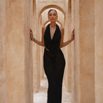 Model wearing a black halter neck dress standing in a sunlit archway with sand-coloured walls.