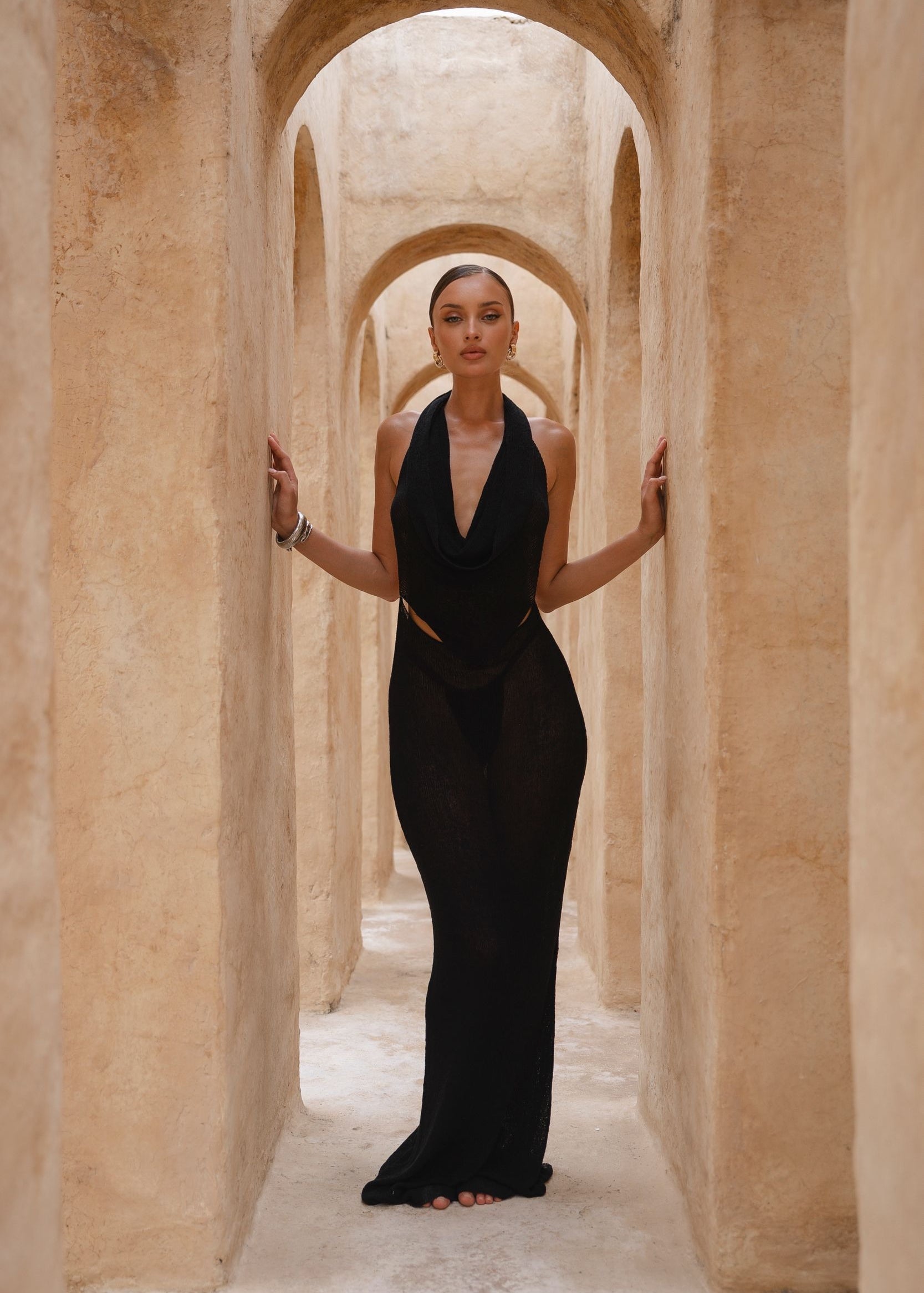 Model wearing a black halter neck dress standing in a sunlit archway with sand-coloured walls.